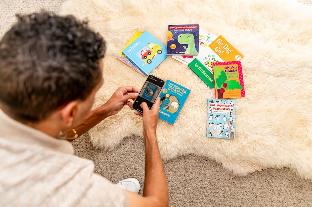 A person photographing a spread of children’s books on a sheepskin rug with their phone.