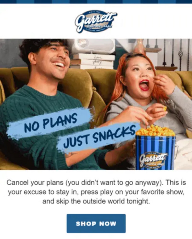Image shows two people on a couch eating popcorn. The person on the left is a tanned-skin person with dark hair, wearing a green sweater. The person on the right is a light-skinned person with orange hair and red lipstick. Both are smiling and eating out of a blue-striped Garrett Popcorn bin.