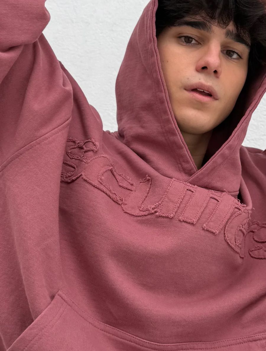 Young man in a dusty rose hoodie with distressed appliqué lettering, looking at the camera.