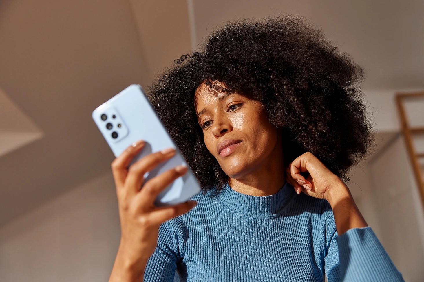 Woman with curly hair looks at a smartphone while sitting indoors in warm sunlight.