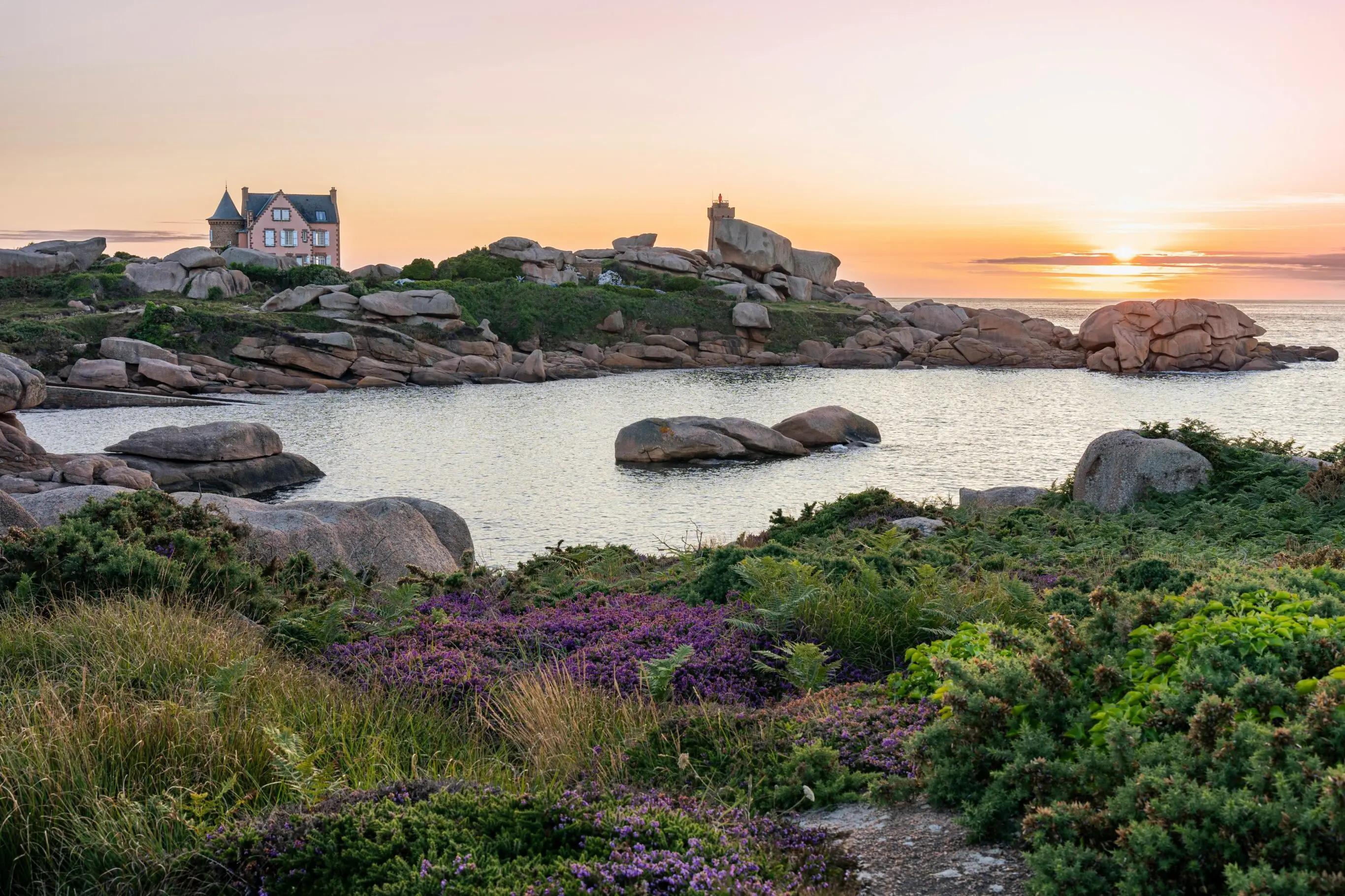 Paysage côtier de la Côte de Granit Rose en Bretagne au coucher du soleil, avec une maison en pierre entourée de rochers sculptés par la mer et de végétation sauvage.