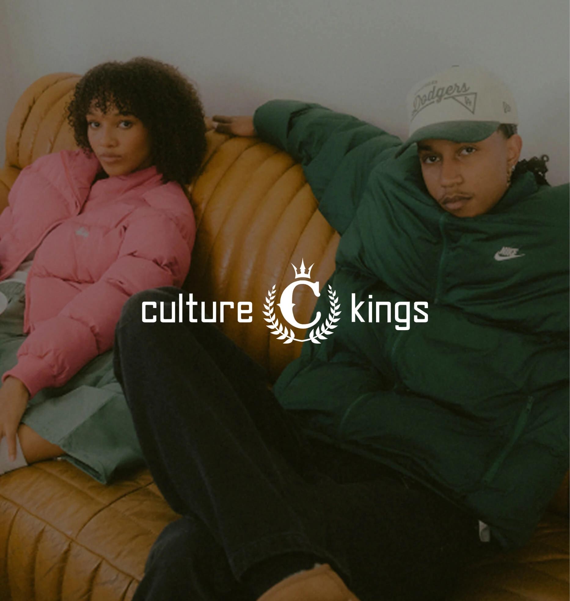 Two people in puffer jackets lounge on a brown leather sofa beneath the Culture Kings logo.