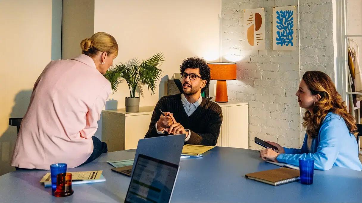Three coworkers discuss work around a table with laptops and notebooks in a colorful office.