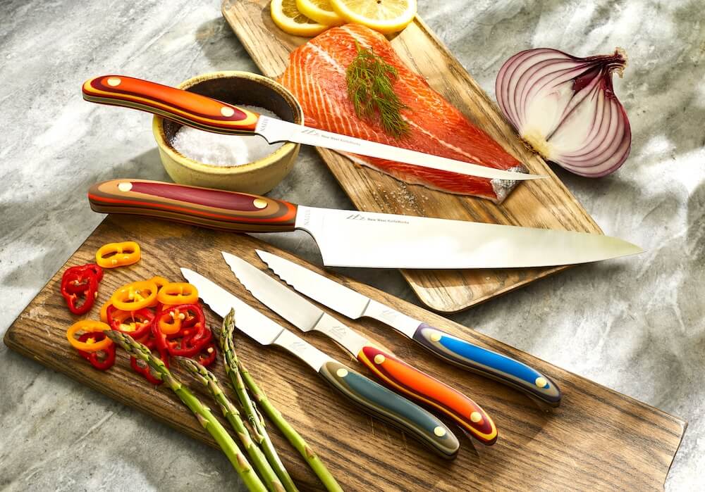 Colorful kitchen knives on wooden boards with salmon, lemon slices, red onion, sliced peppers, and asparagus.