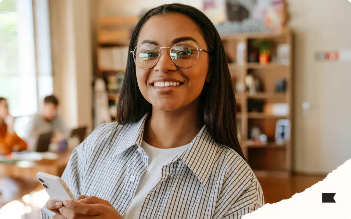 Smiling person with glasses holding a smartphone, standing in a room with bookshelves and people in the background.