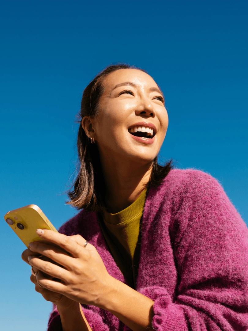 Smiling woman in a purple sweater holds a yellow smartphone against a bright blue sky.