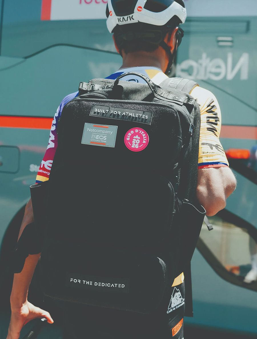 A cyclist from behind, wearing a helmet and a jersey, carries a black backpack with Giro d'Italia and Ineos Cycling Team patches.