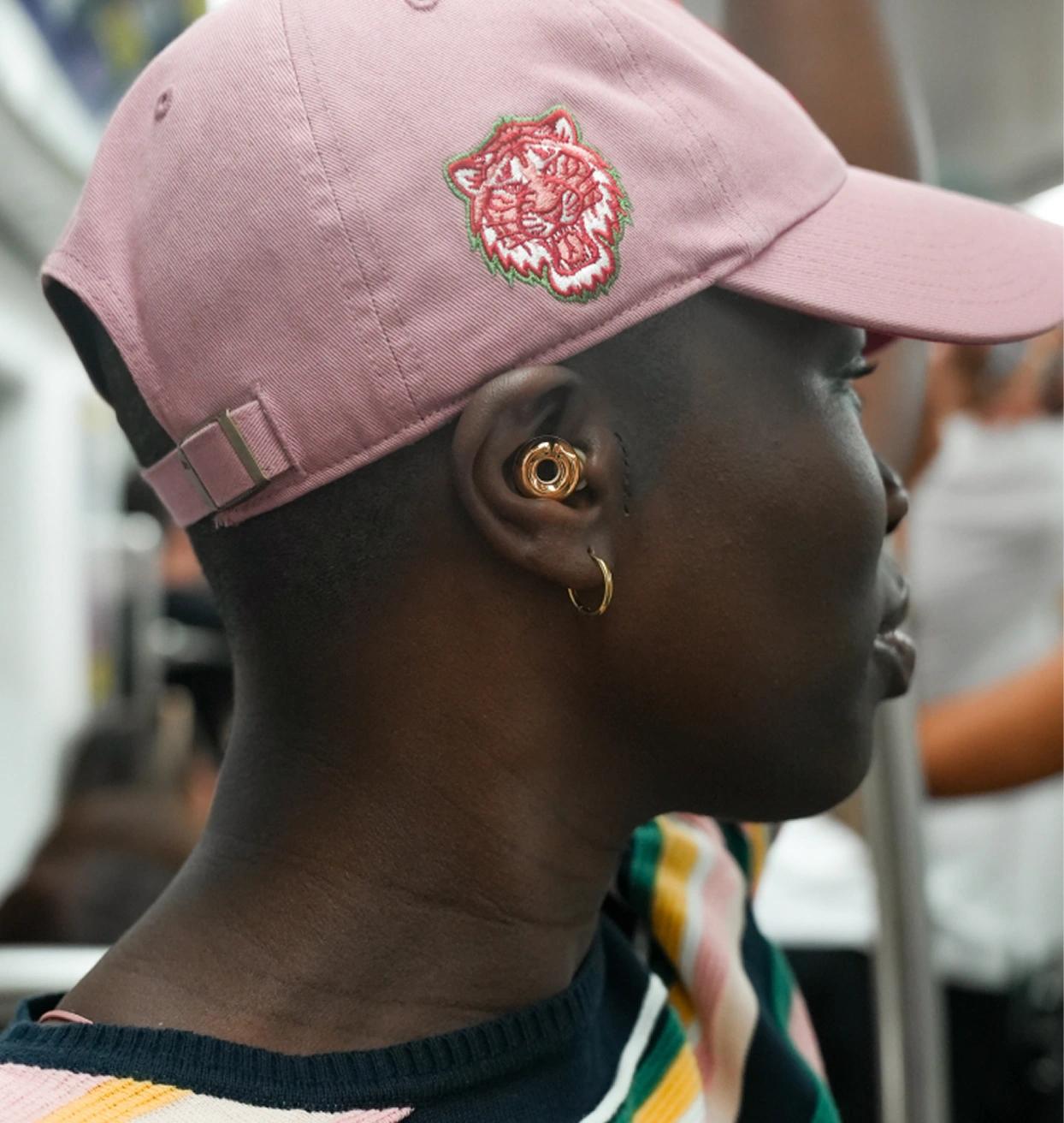Person in profile wearing a pink baseball cap with an embroidered tiger and gold ear jewelry