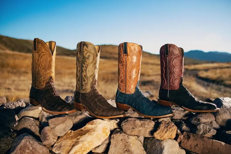 Four colorful cowboy boots with exotic leather and embroidered shafts on a stone wall.