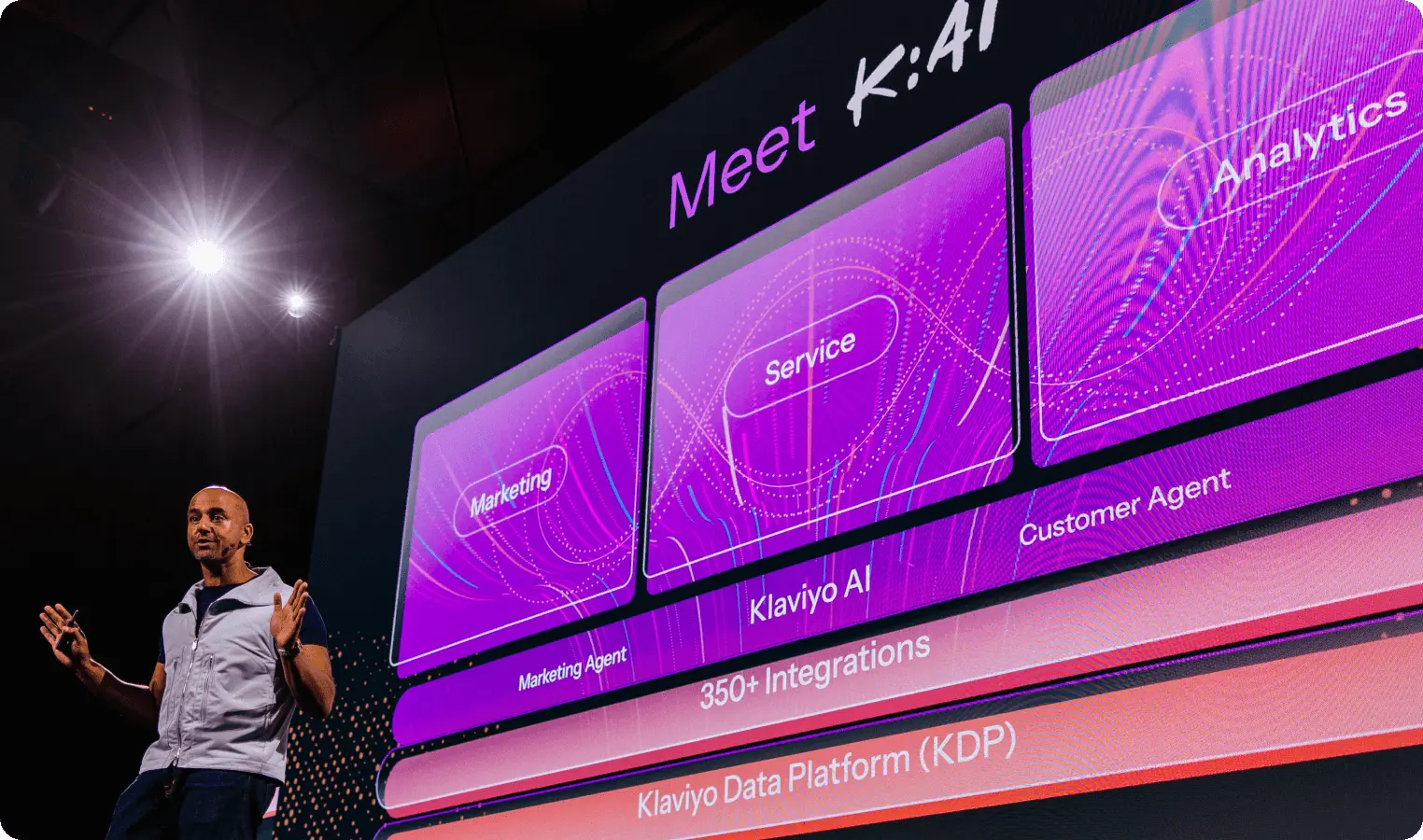 A person presenting in front of a large screen displaying "Meet K:AI" with sections labeled Marketing, Service, and Analytics.