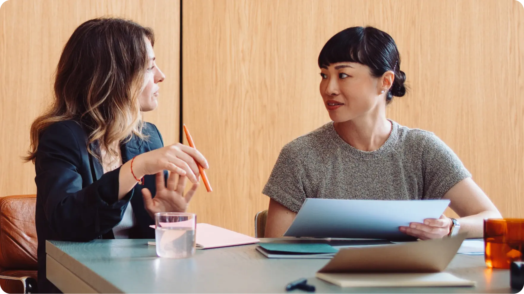 Two women sitting at a table, chatting while holding documents and a pen.