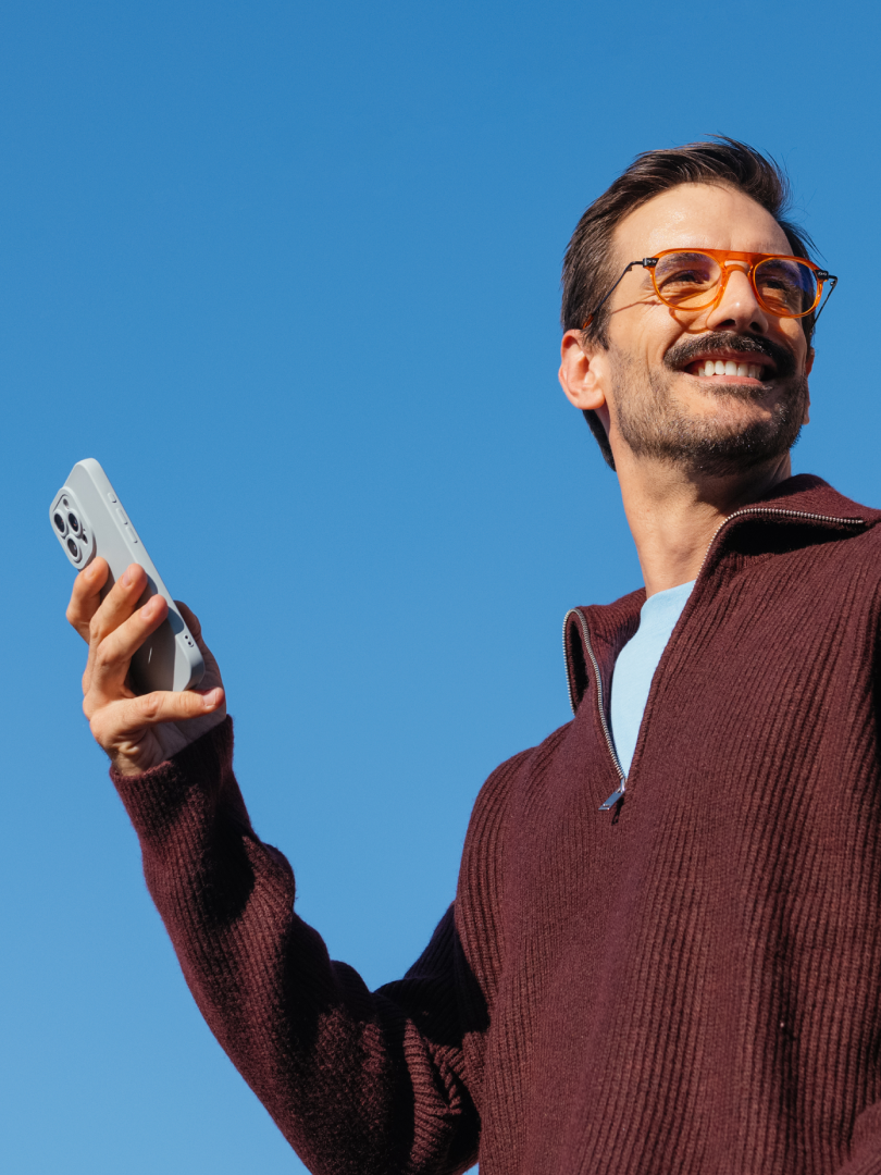 Smiling man in orange glasses holds a smartphone against a clear blue sky.