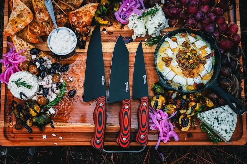 A large charcuterie board laden with cheeses, crackers, grapes, roasted vegetables, a pan of baked brie, and three chef's knives with red handles.