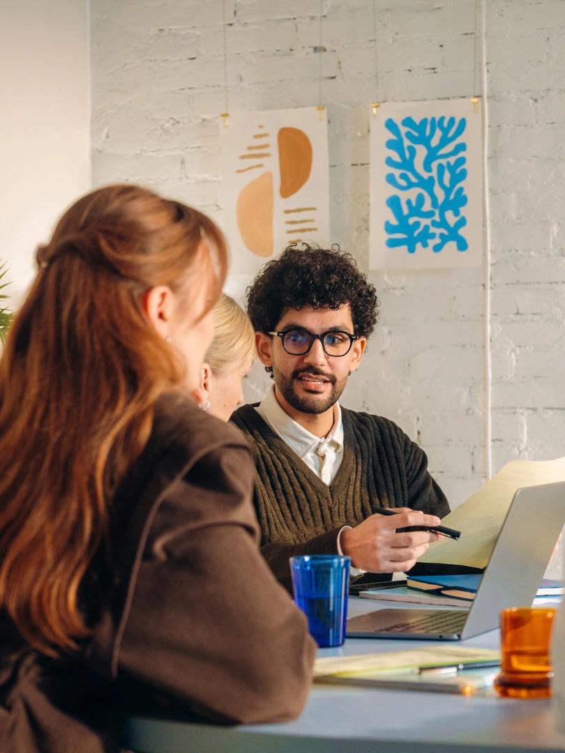 Three coworkers discuss documents around a table with a laptop in a creative office.