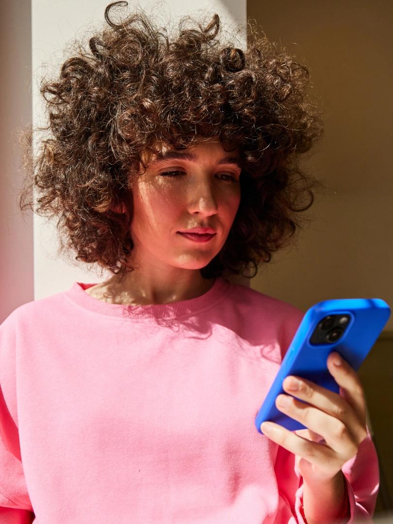 Person with curly brown hair in a pink sweatshirt looking at a blue smartphone.