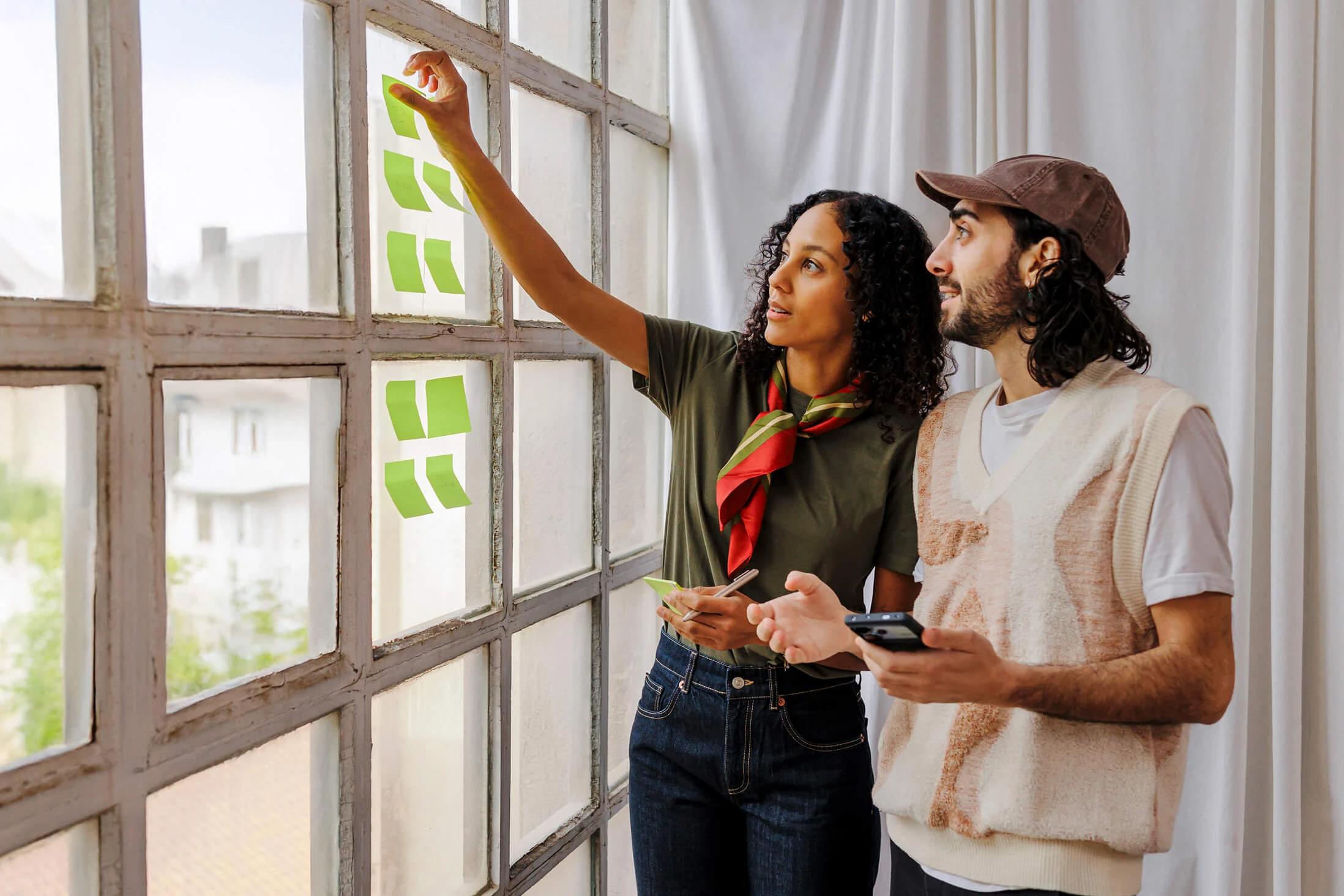 Two coworkers arrange green sticky notes on a large window while holding smartphones.