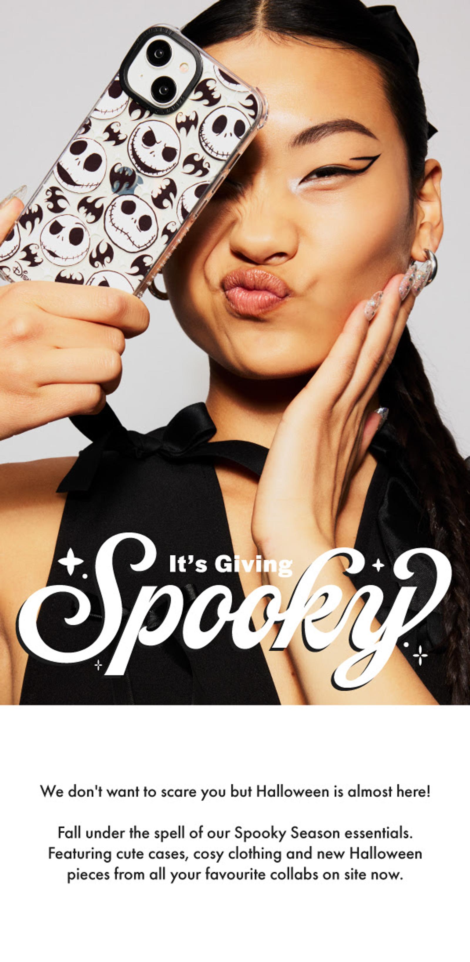 A young woman with winged eyeliner and a black hair bow makes a puckered face, holding an iPhone with a Jack Skellington case over her eye. White graphic text reads "It's Giving Spooky". Below, smaller text promotes Halloween cases, clothing, and collaborations.
