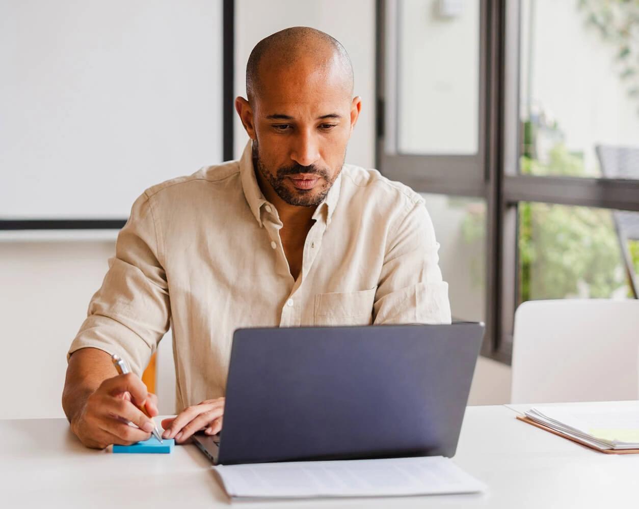 Man takes notes while working on a laptop at a bright office desk.