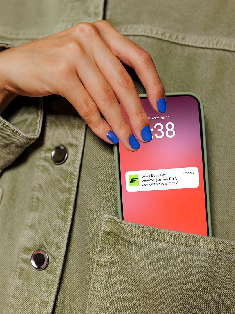 Hand with blue nail polish pulls a smartphone showing a saved-item notification from a green jacket pocket.