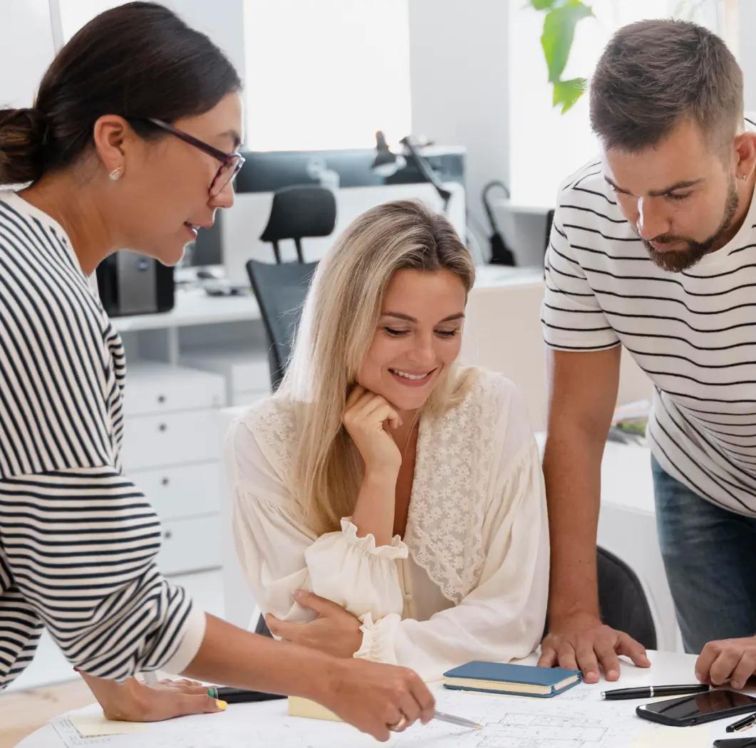 Three people collaborating around a table, looking at documents and discussing ideas in an office setting.