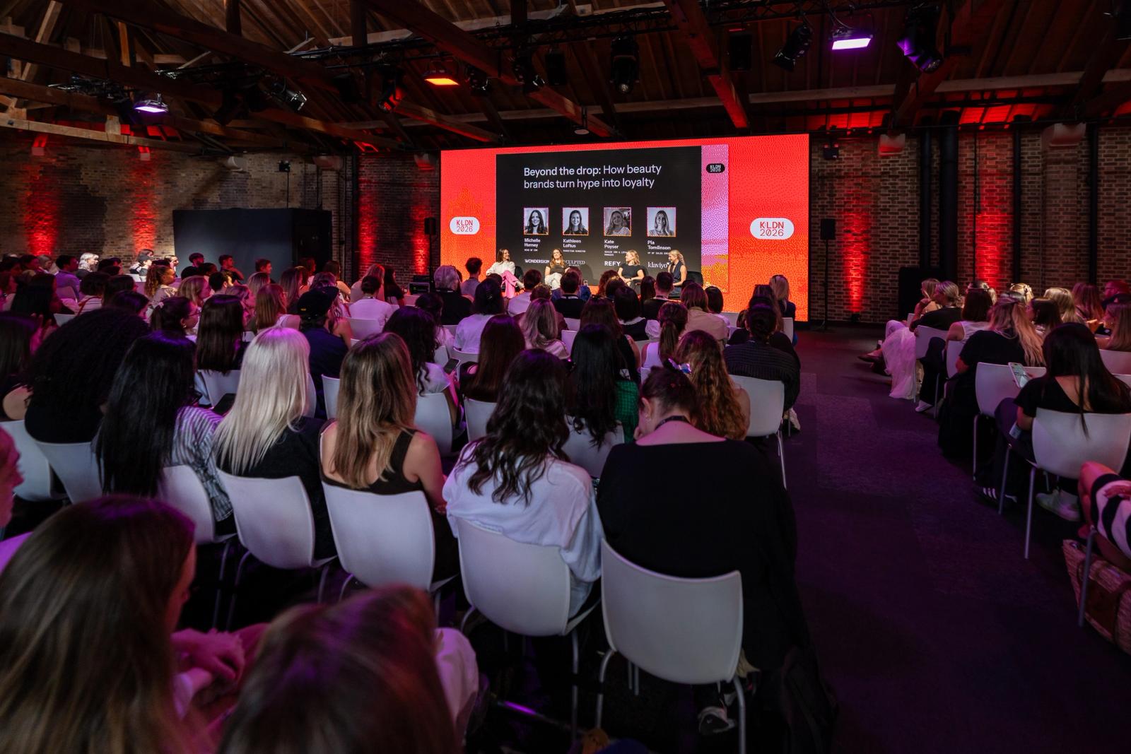 Audience watches a panel discussion titled 'Beyond the drop: How beauty brands turn hype into loyalty' on a large screen at an event.