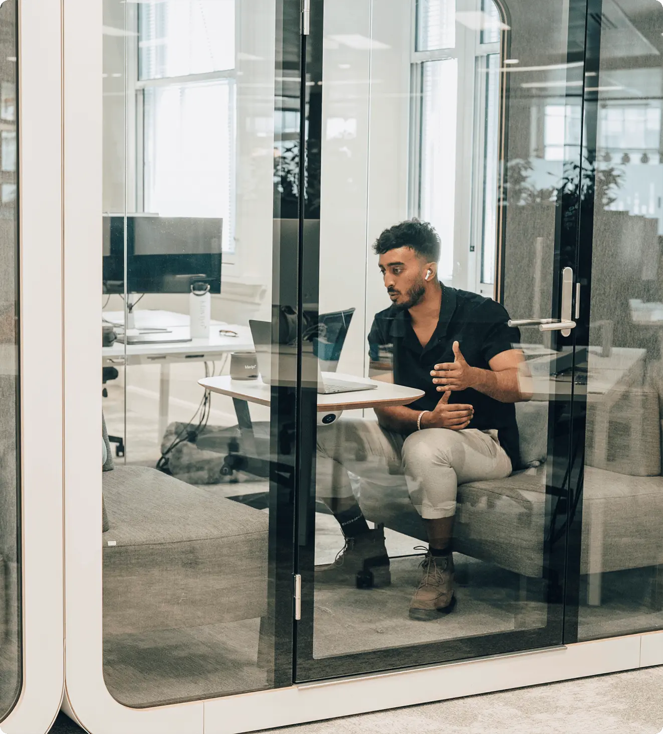 Person in a glass office booth, wearing earbuds, gesturing while on a laptop video call.