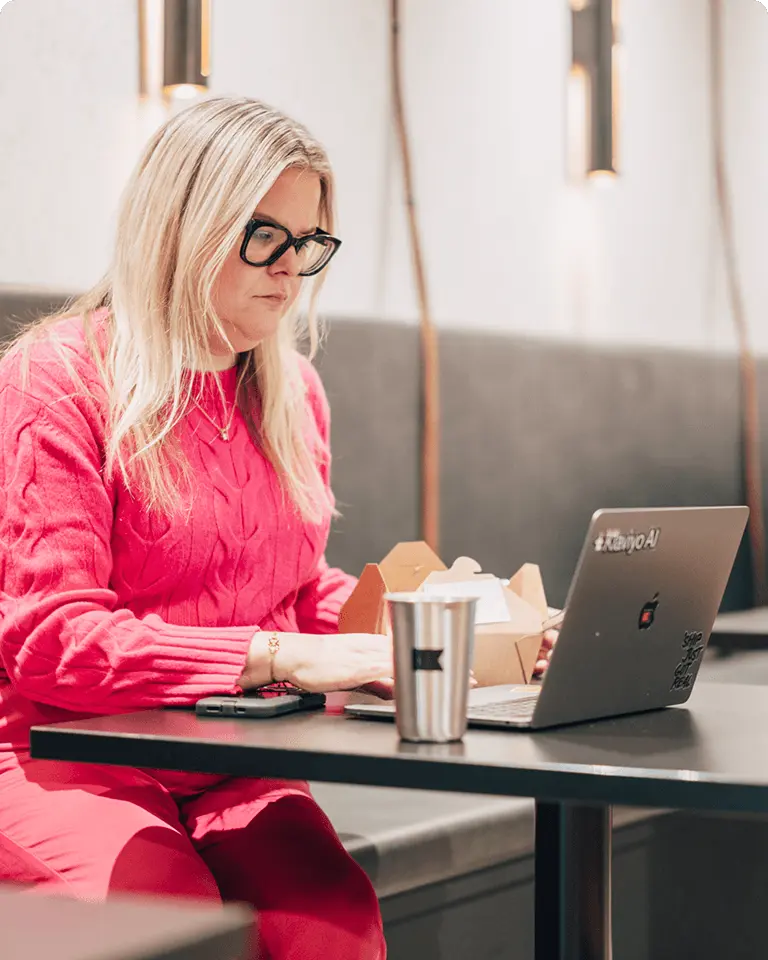 Person in a pink sweater working on a laptop at a table, with a takeout box and a metal cup nearby.