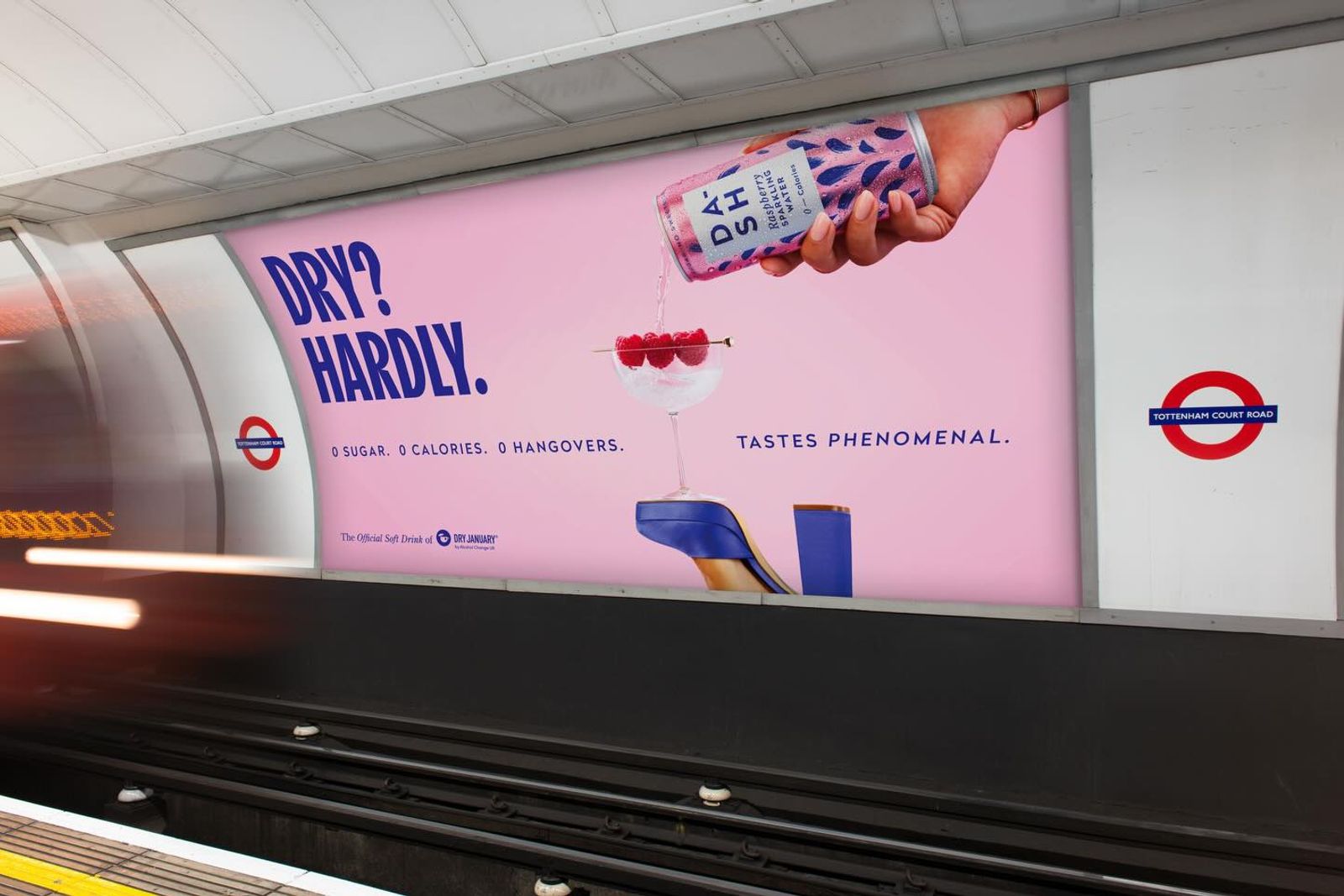 A large pink advertisement on a subway platform shows a hand pouring a canned drink into a cocktail glass with raspberries. Bold text reads “DRY? HARDLY.” with smaller text saying “0 sugar. 0 calories. 0 hangovers. Tastes phenomenal.” The setting appears to be an underground train station with tracks in the foreground.