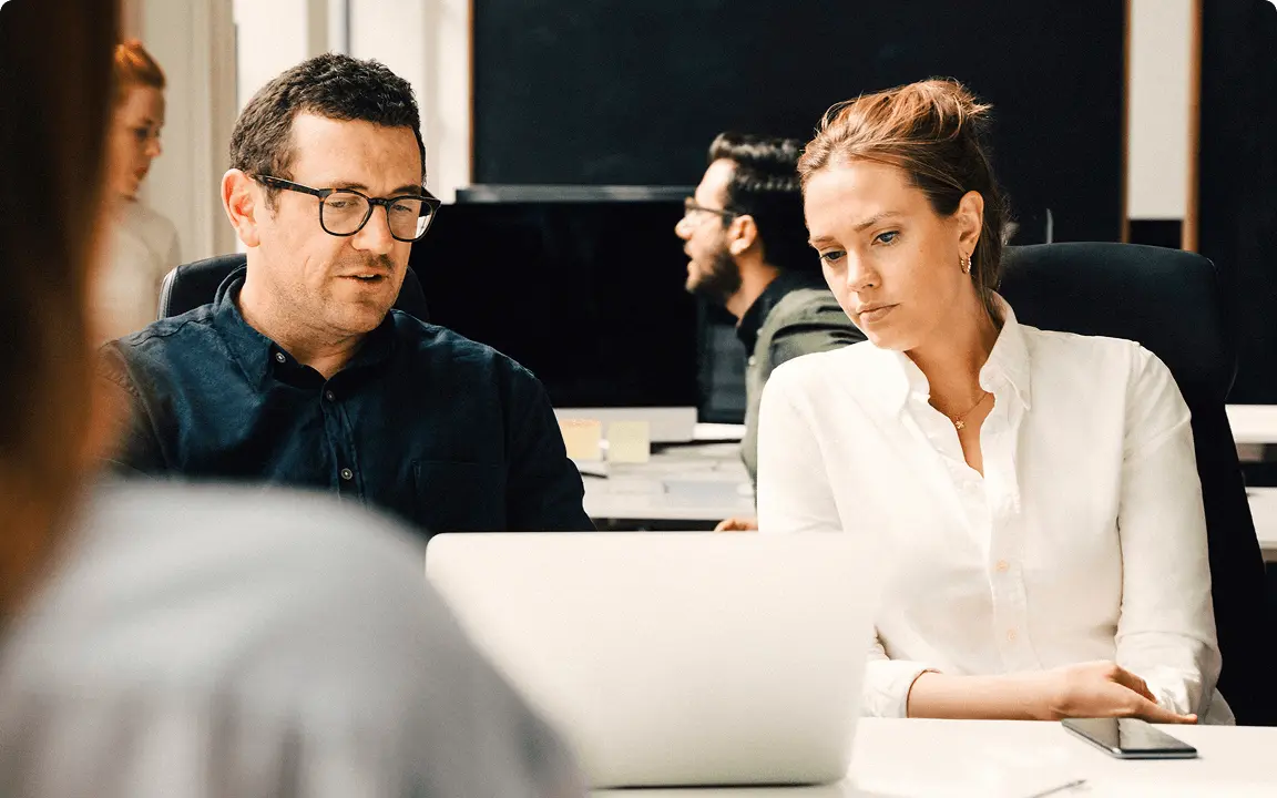 Two people sit at a desk in an office, looking at a laptop screen together. Other coworkers are visible in the background, working and talking.