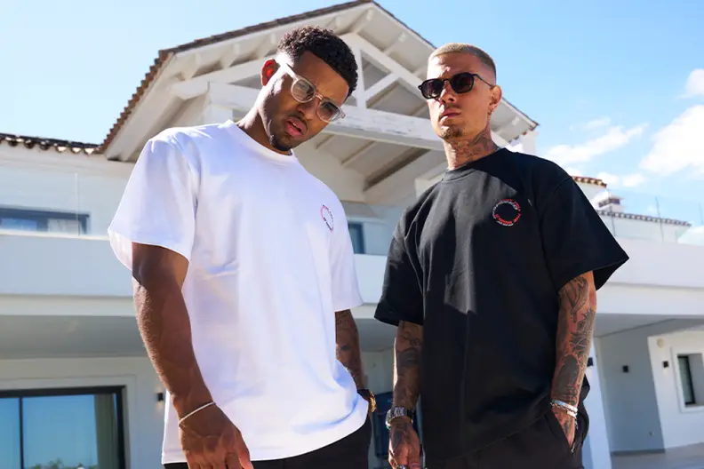 Two men wearing white and black logo t-shirts and sunglasses pose in front of a white building.