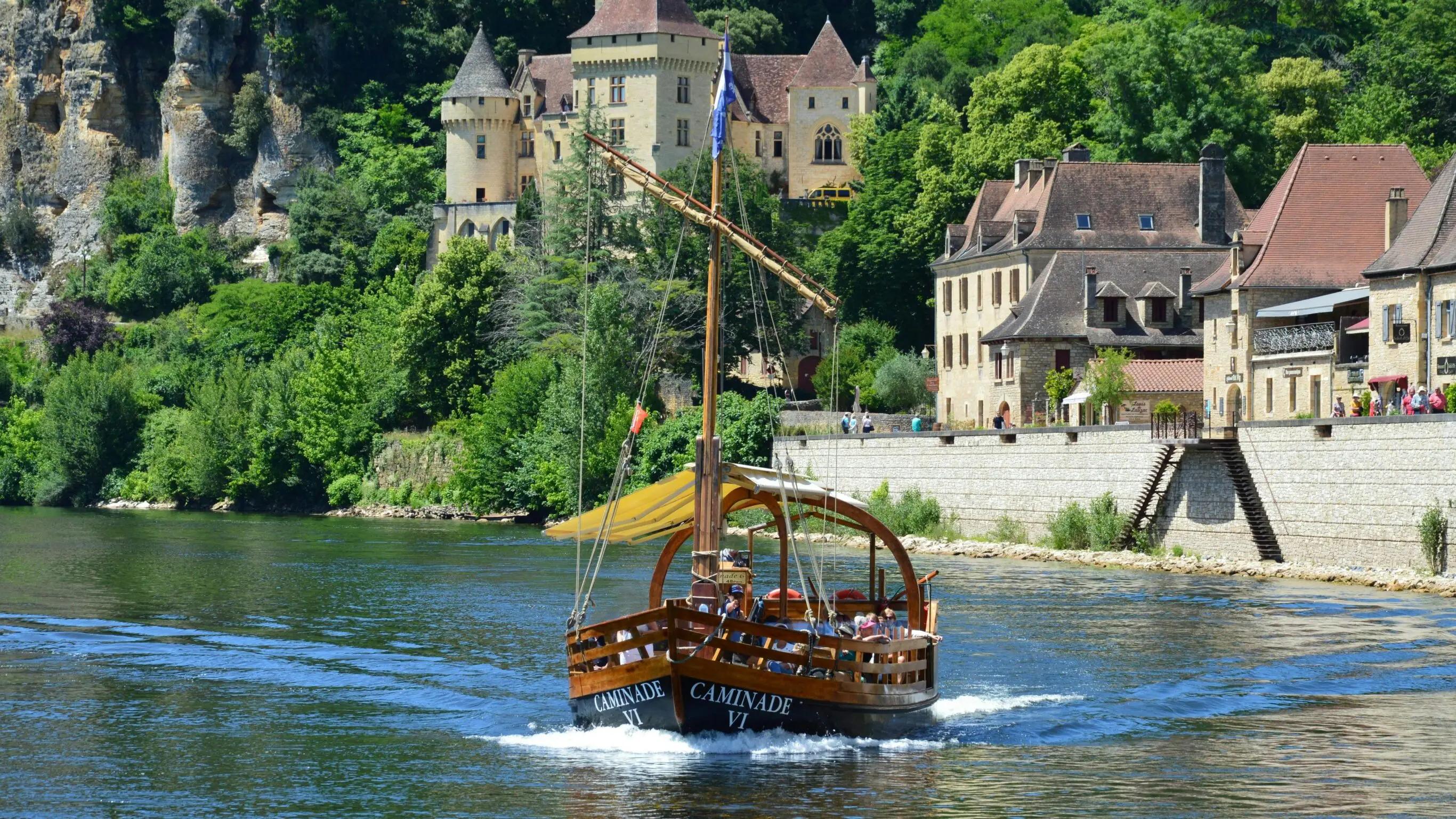 Une gabare traditionnelle naviguant sur la Dordogne, avec en arrière-plan un château médiéval et des maisons en pierre typiques du Périgord noir.