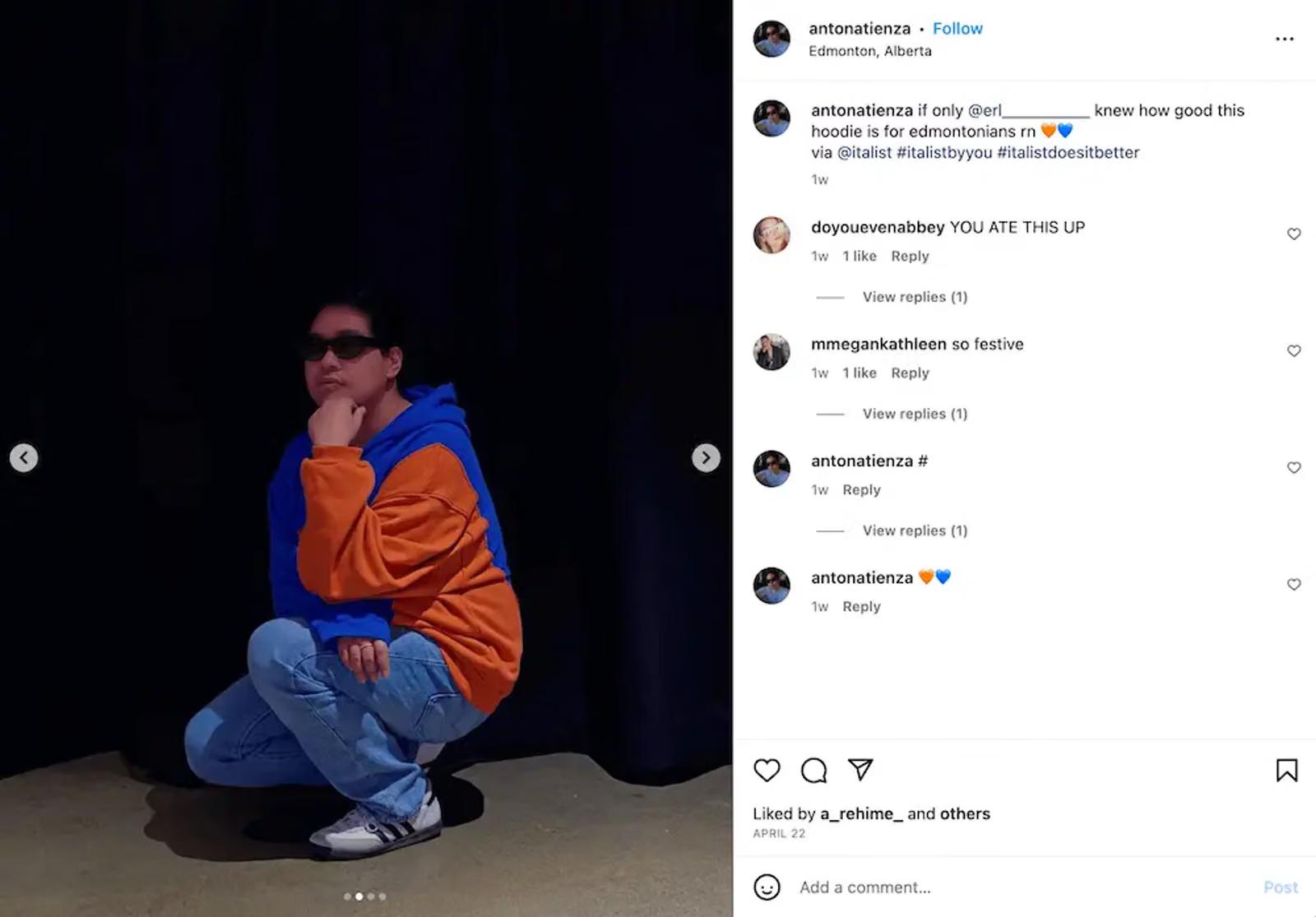 A person wearing sunglasses and a blue and orange hoodie squats with their hand on their chin against a dark background.
