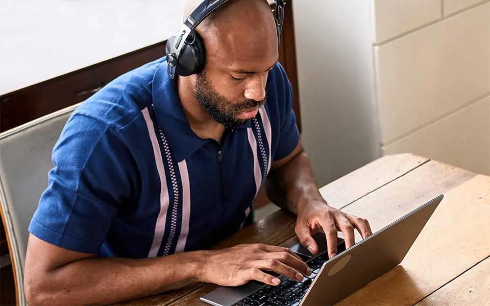 Uomo con cuffie lavora al laptop seduto a un tavolo di legno.