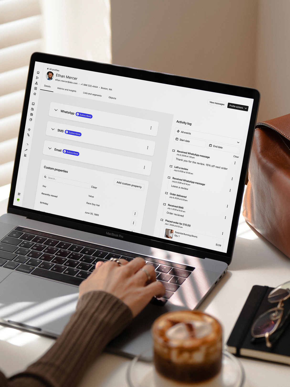 Person using a laptop displaying a customer profile and activity log beside an iced coffee.