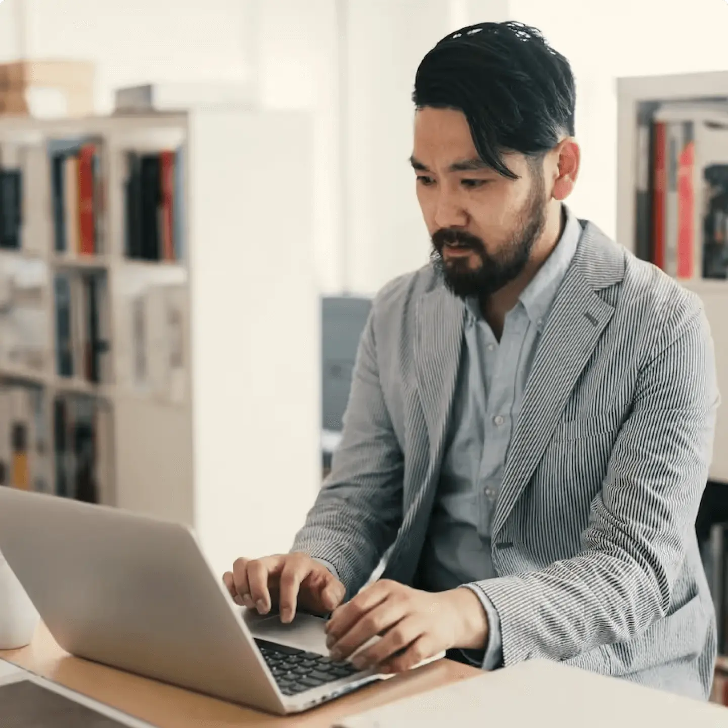 Man in a striped blazer working on a laptop at a desk in an office setting.
