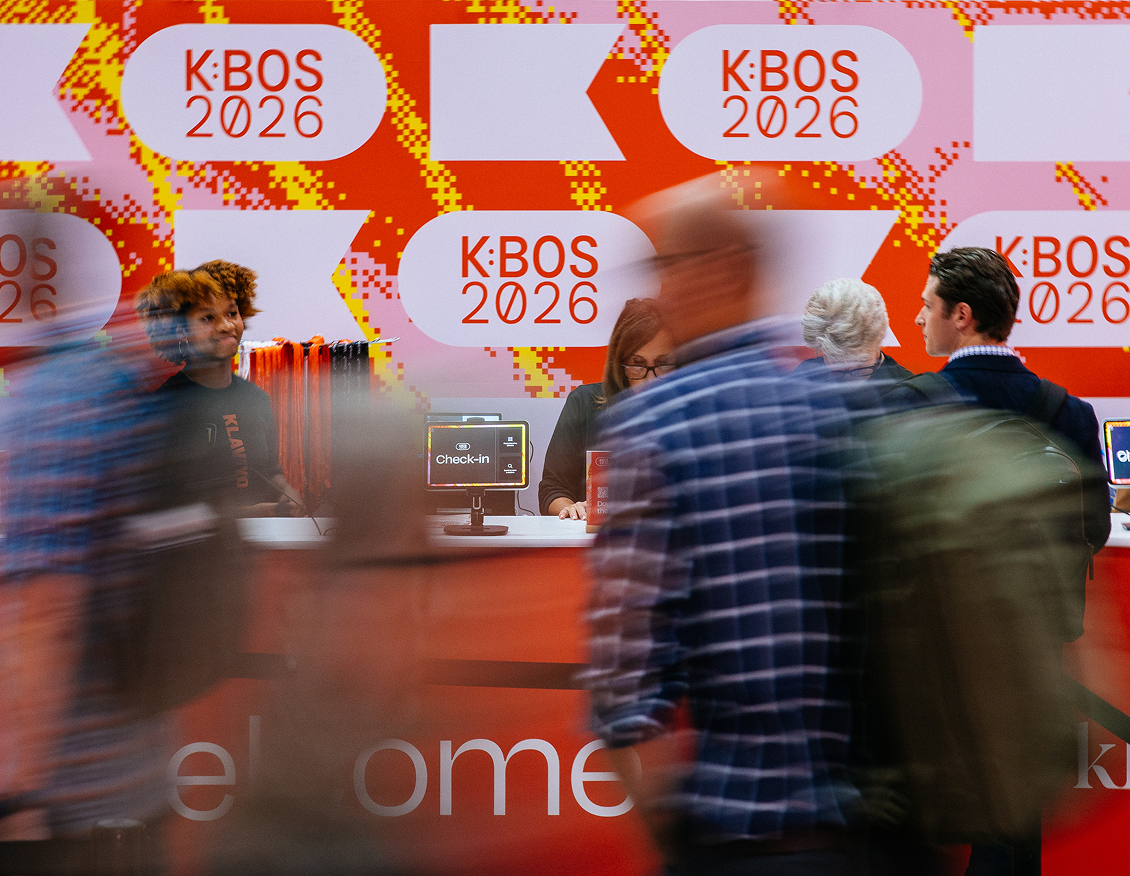 Attendees check in at a busy K:Bos 2026 registration desk as people pass in the foreground.