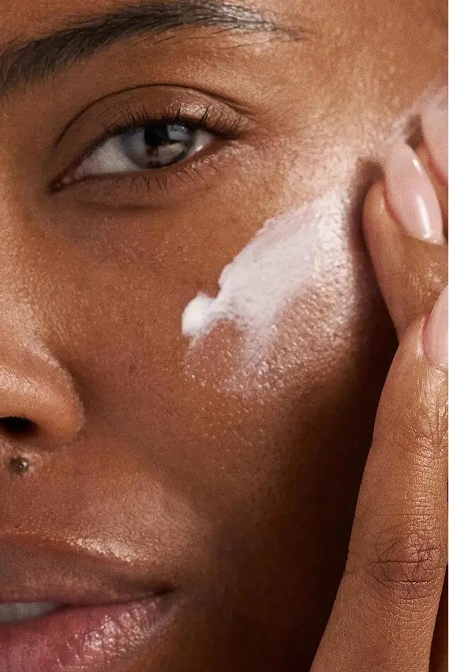 Close-up of a person applying white skincare cream to their cheek with two fingers.