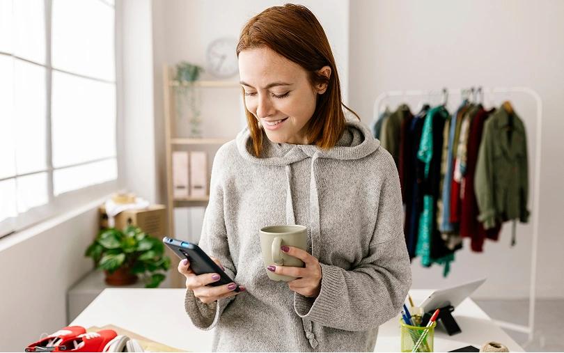 Smiling woman checks her smartphone while holding a mug in a bright clothing studio.