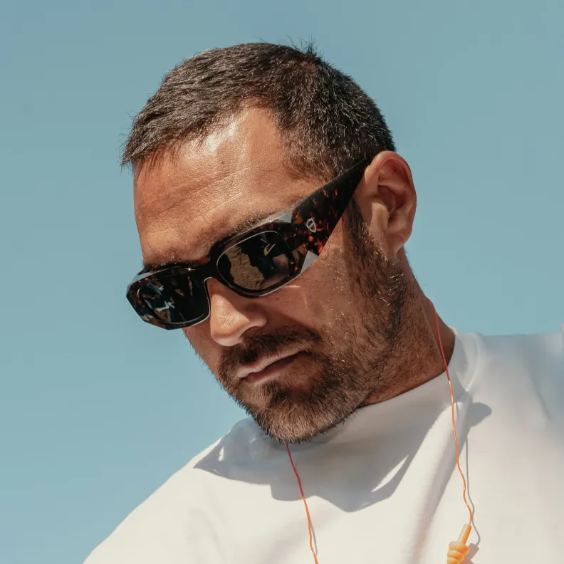 Man in tortoiseshell sunglasses with orange earplugs around his neck, looking down under a blue sky.