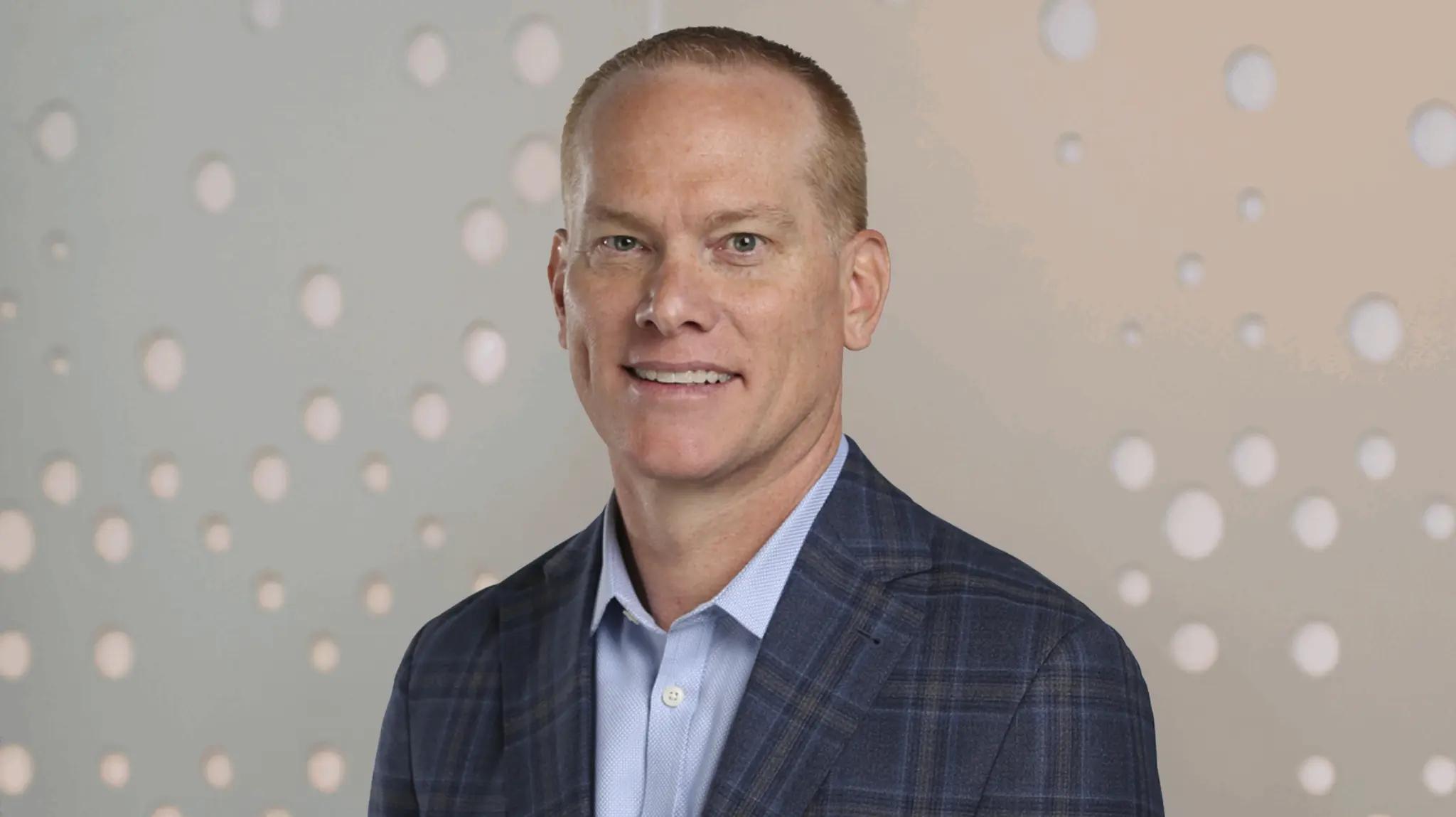 Smiling man in a blue plaid suit jacket and light blue shirt, standing against a patterned background.