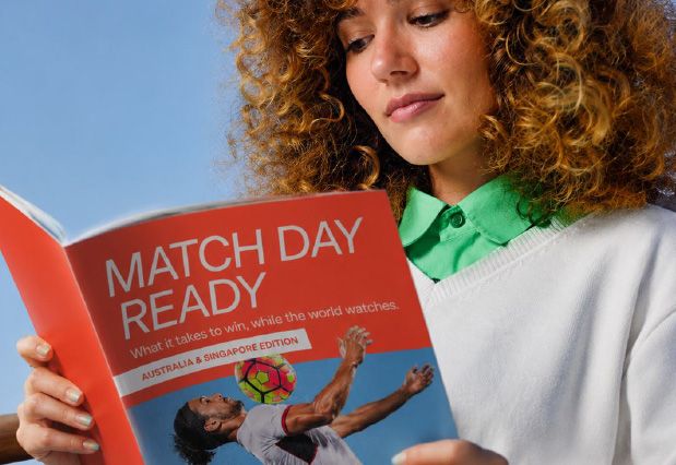 A woman with curly hair reads a magazine titled "MATCH DAY READY" which features a soccer player heading a colorful ball.