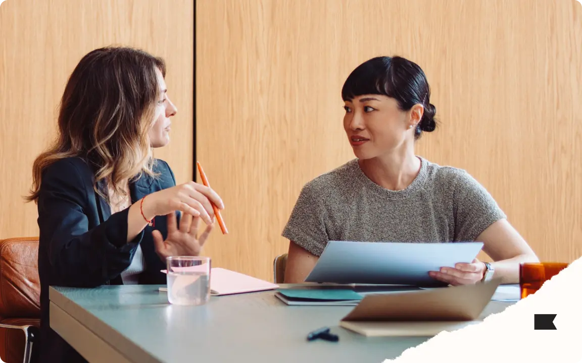 Two women sitting at a table, engaged in conversation, one holding a pen and the other holding papers.