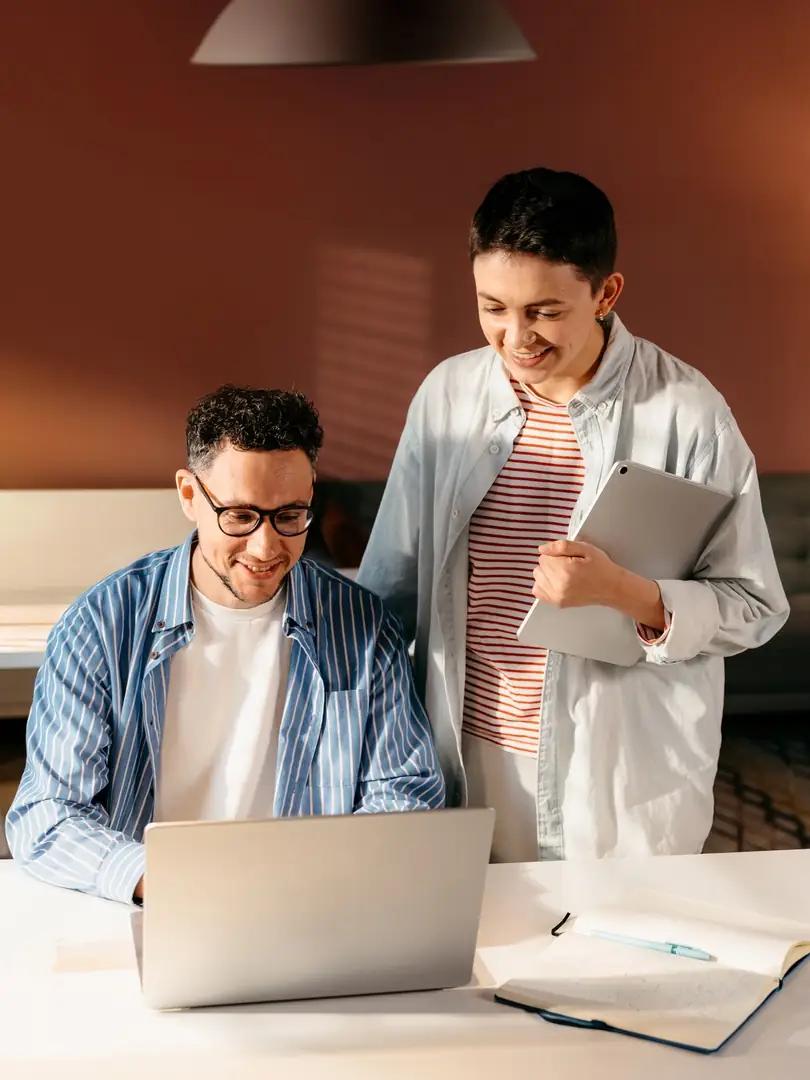 Two smiling coworkers review a laptop together while one holds a tablet.