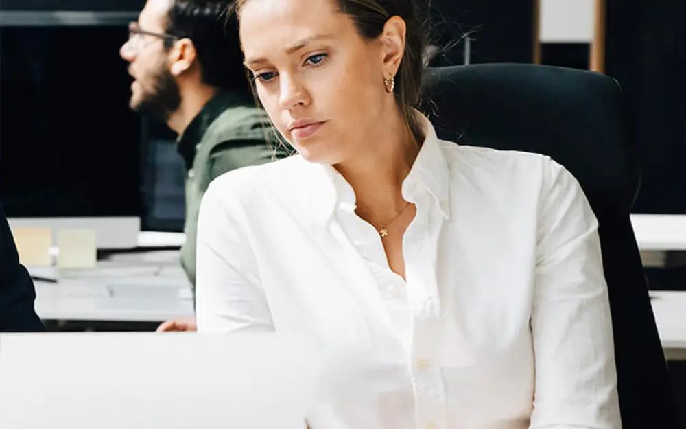Frau in weißem Hemd arbeitet konzentriert am Computer, im Hintergrund ein weiterer Mann.