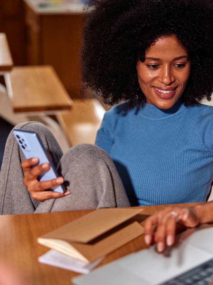 Smiling woman holds a smartphone while using a laptop at a table with notebooks.