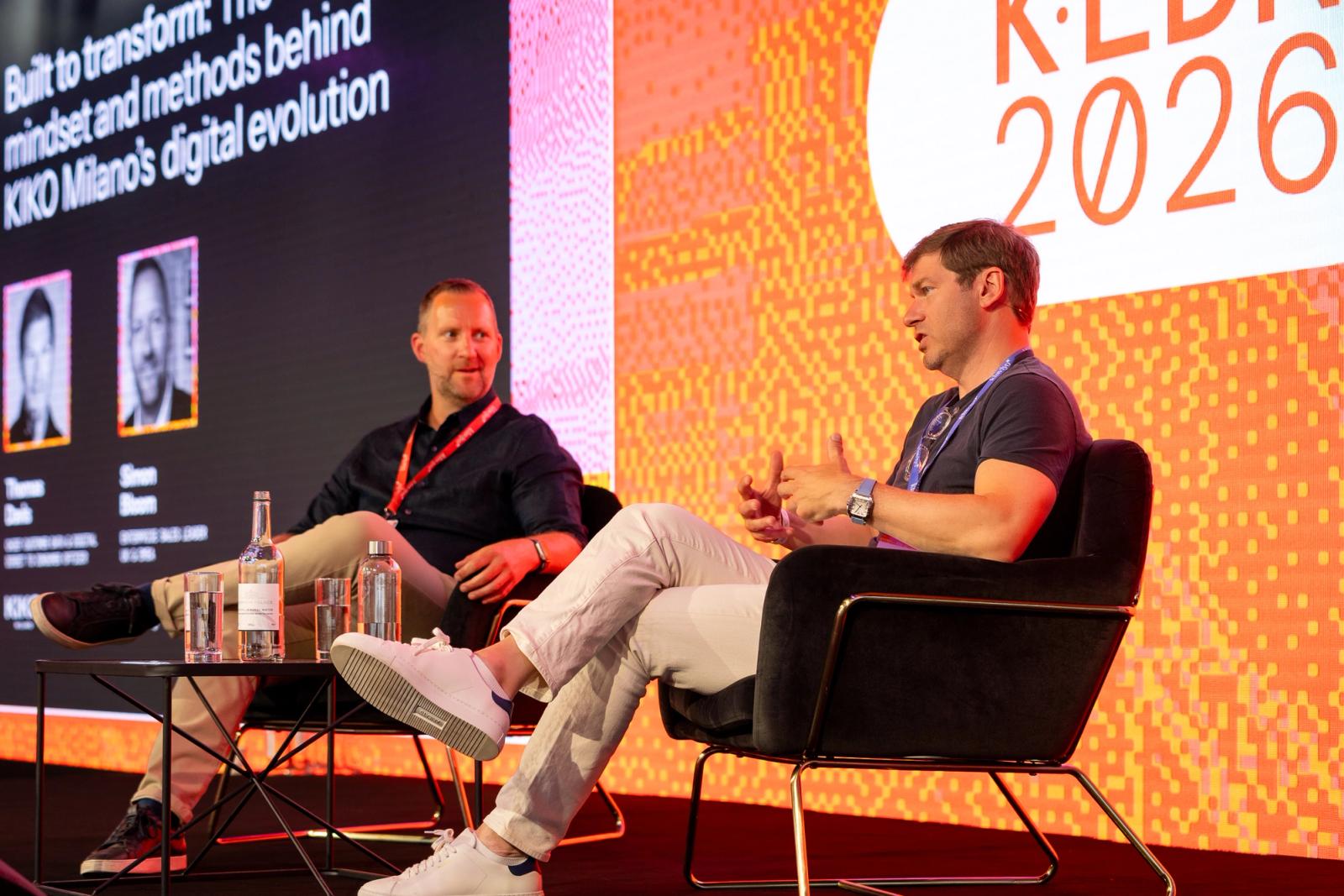 Two men sit on a stage for a panel discussion, with a large screen behind them displaying presentation slides and 'K-LED 2026'.