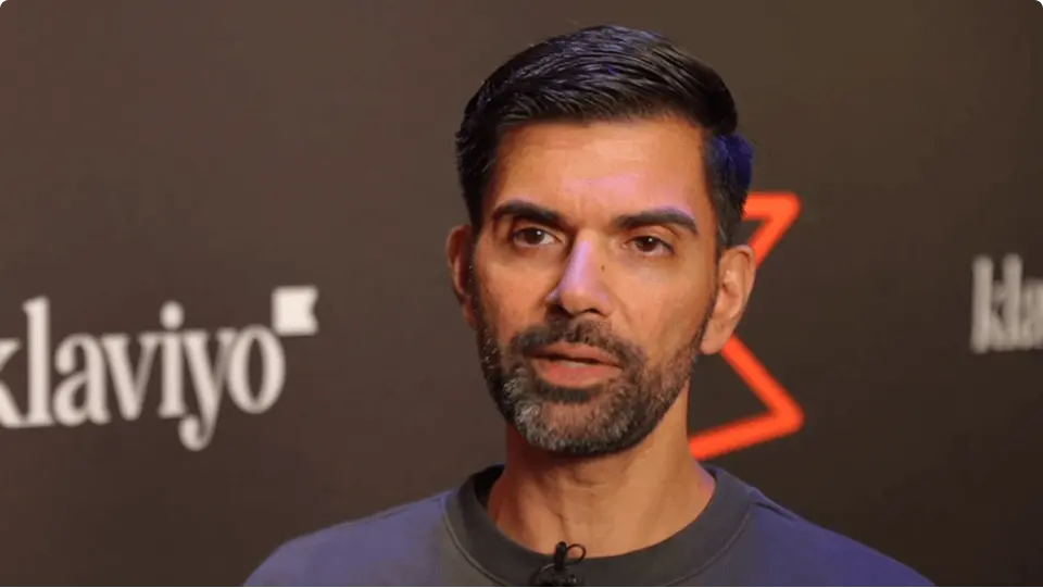 A man with short dark hair and a beard speaks in front of a backdrop featuring the Klaviyo logo.