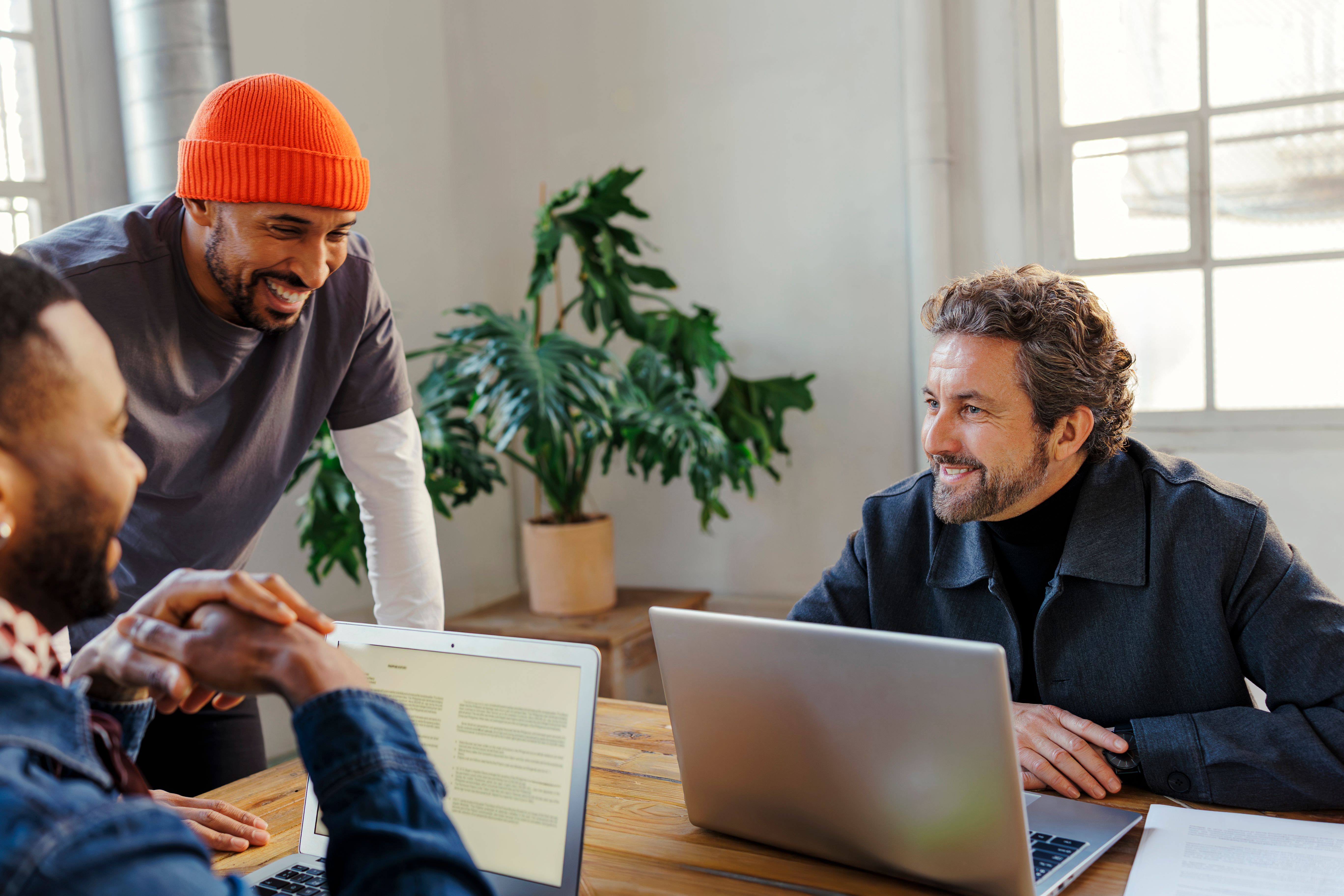 Three smiling men collaborating at a table with laptops, two shaking hands.