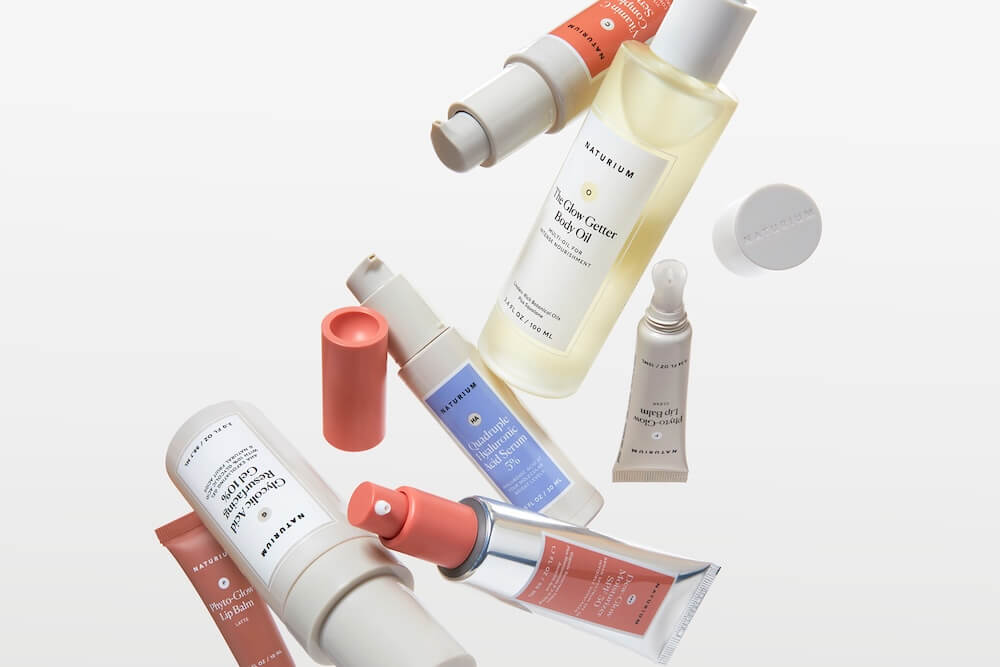 Naturium skincare products, including body oil, serum, and lip balm, arranged diagonally on a white background.