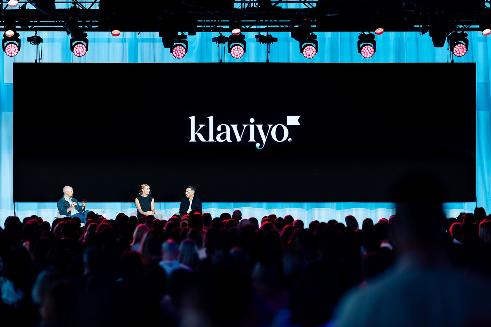 A man in a dark jacket speaks on a stage, gesturing towards a screen that reads "CHOOSE CONFLICTING GOALS".